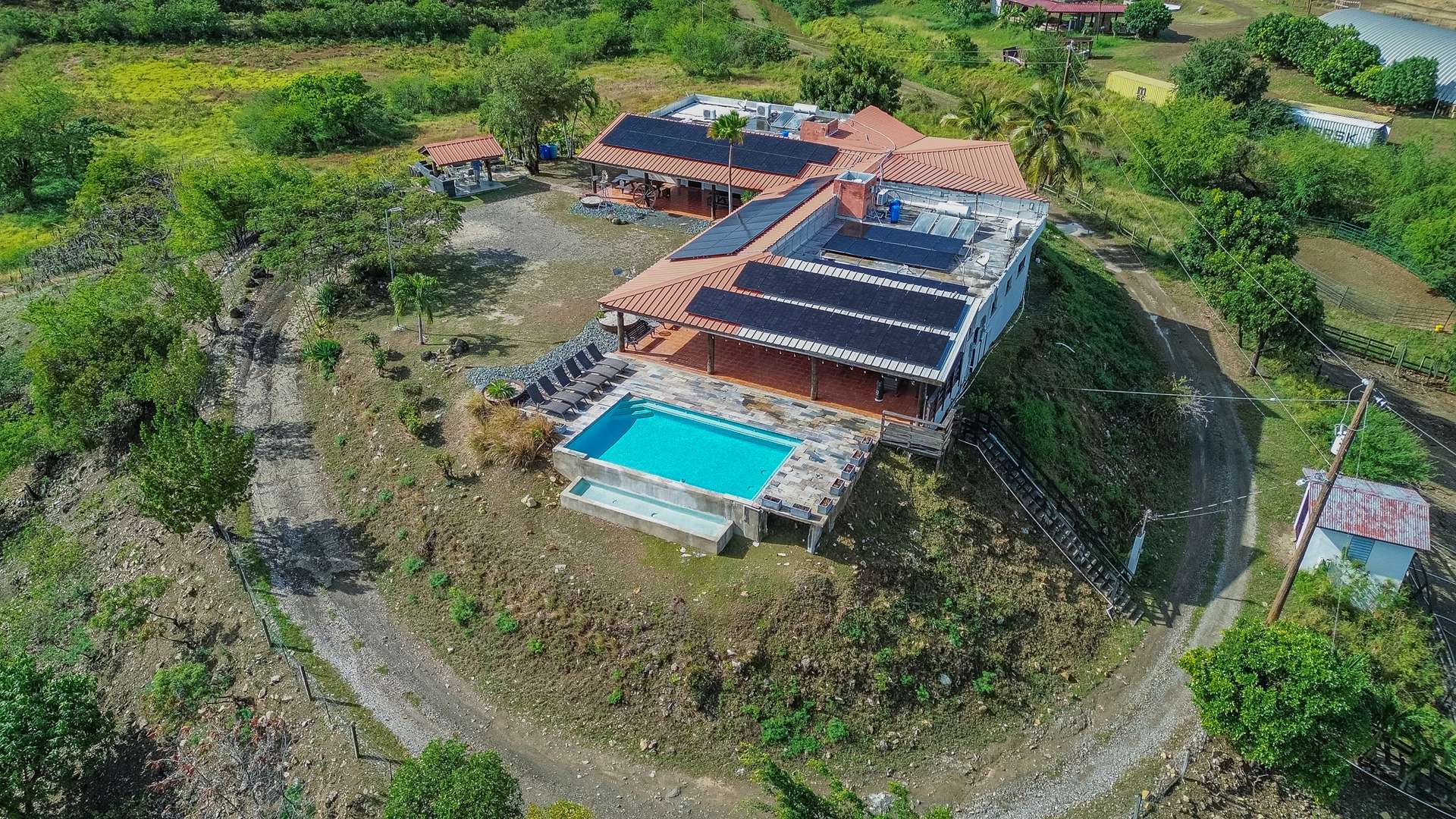 Overhead drone view of the hilltop house, its solar-panelled roof, the swimming pool terrace and the gravel track winding up through green scrub