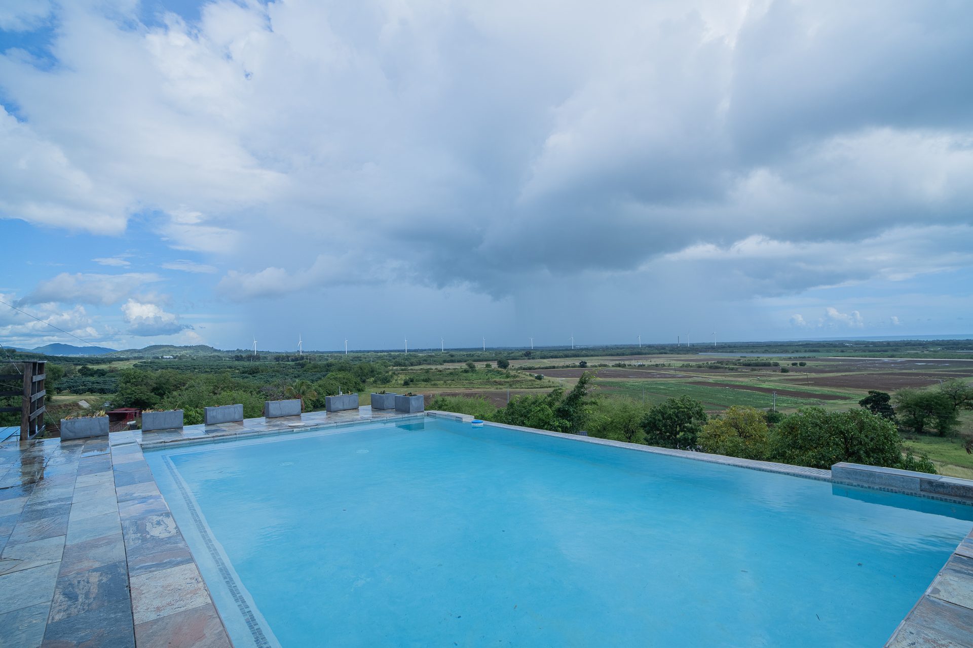 The infinity pool edge dissolving into the farmland valley below, with towering clouds overhead