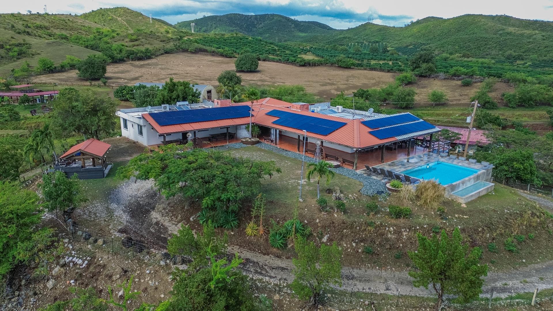 Drone view of the long red-roofed ranch house and pool from the side, with rolling green hills and farmland stretching behind it