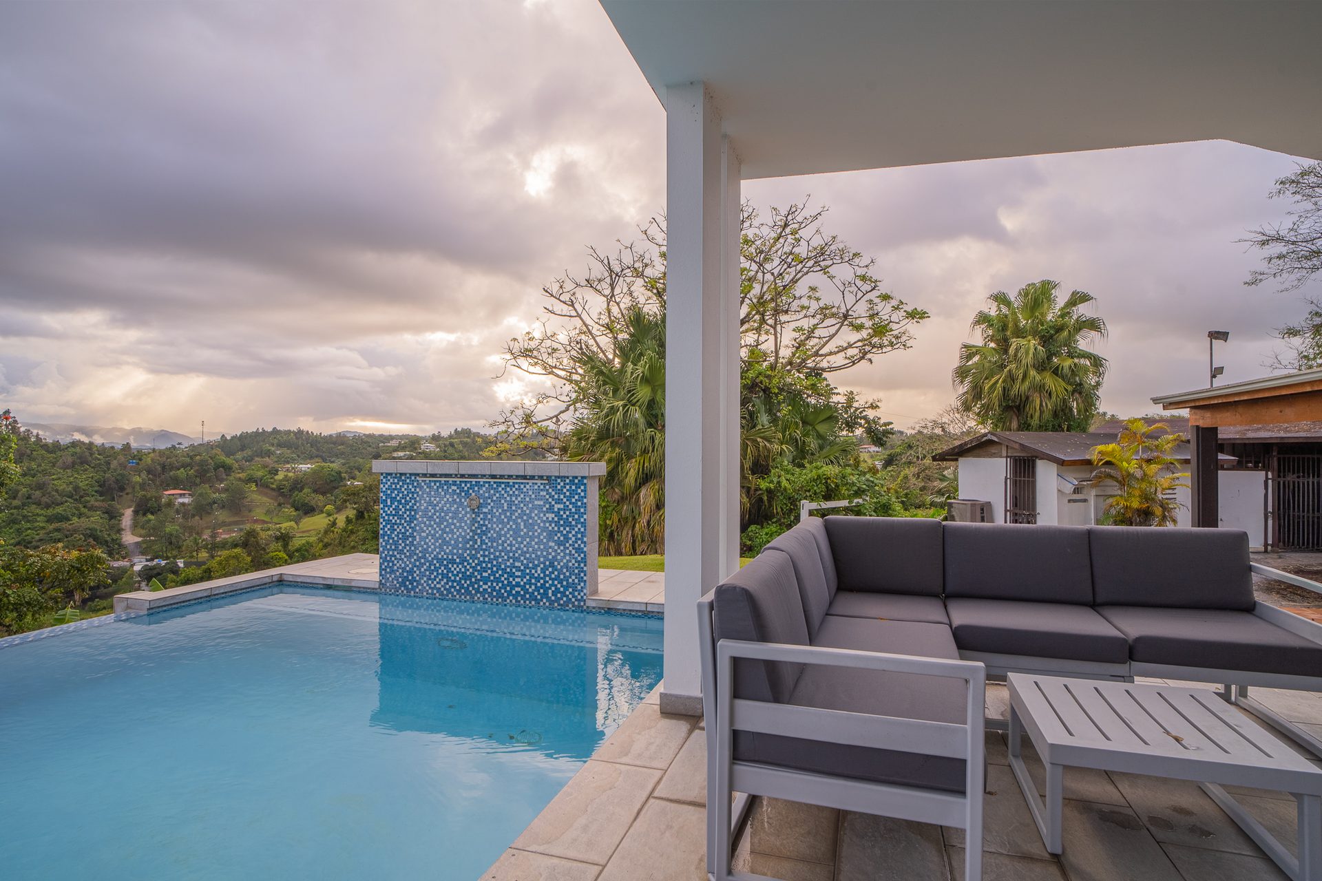 The infinity pool and grey outdoor lounge seen at dusk as the sky glows over the Cayey hills